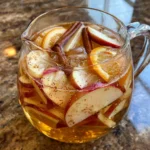 Pitcher of apple cider sangria with apple slices, orange slices, and cinnamon sticks, served in glasses over ice.